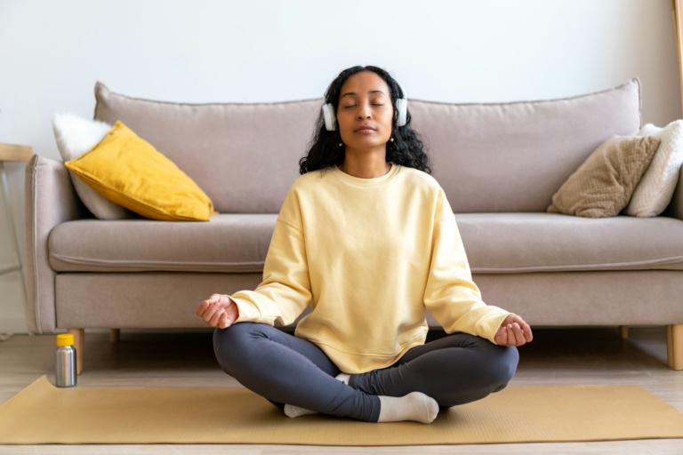 young african american female in headphone listening to music while meditating on mat in living room
